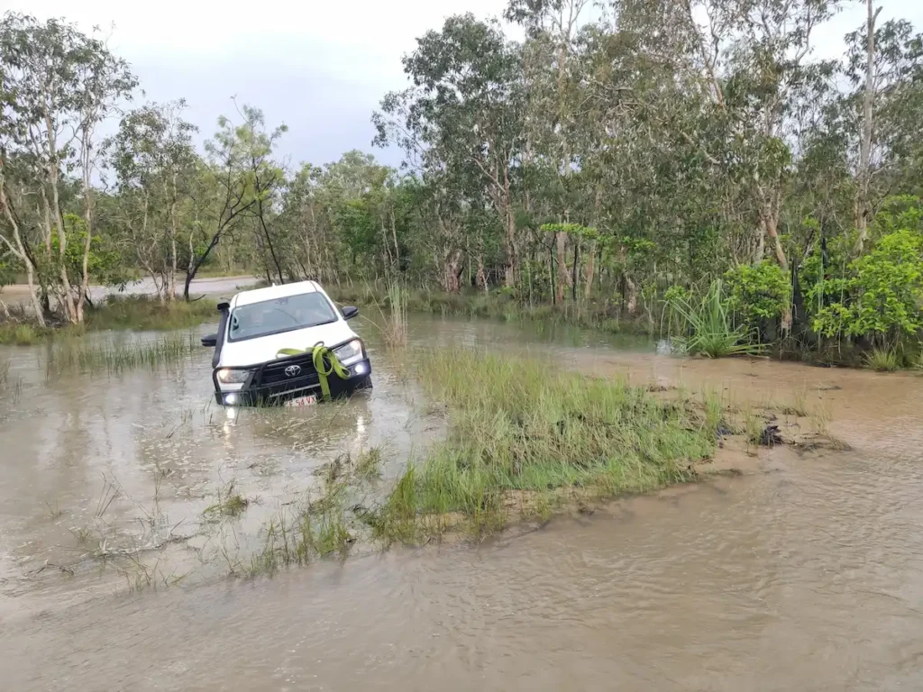 A white Toyota Hilux 4WD vehicle driving through a deep water crossing on a flooded dirt road in the Northern Territory.