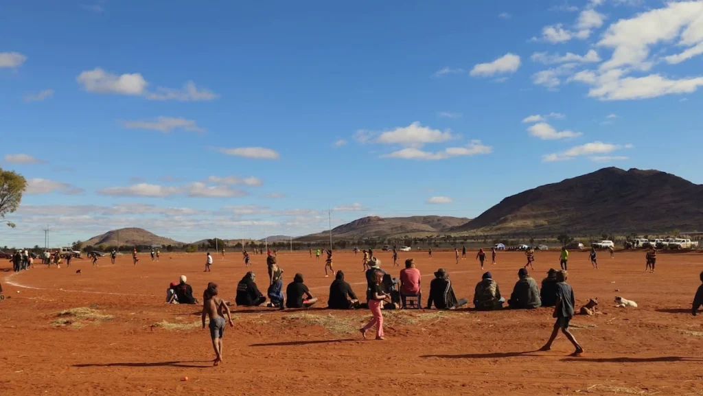 Local community gathering for a football match in the red desert of the Australian Outback with mountains in the background.
