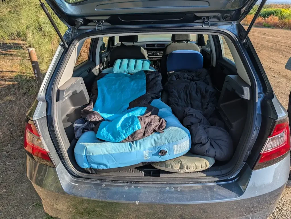 The open trunk of a small hatchback car showing a travel sleeping setup with a blue inflatable mattress, pillows, and a sleeping bag for budget camping.