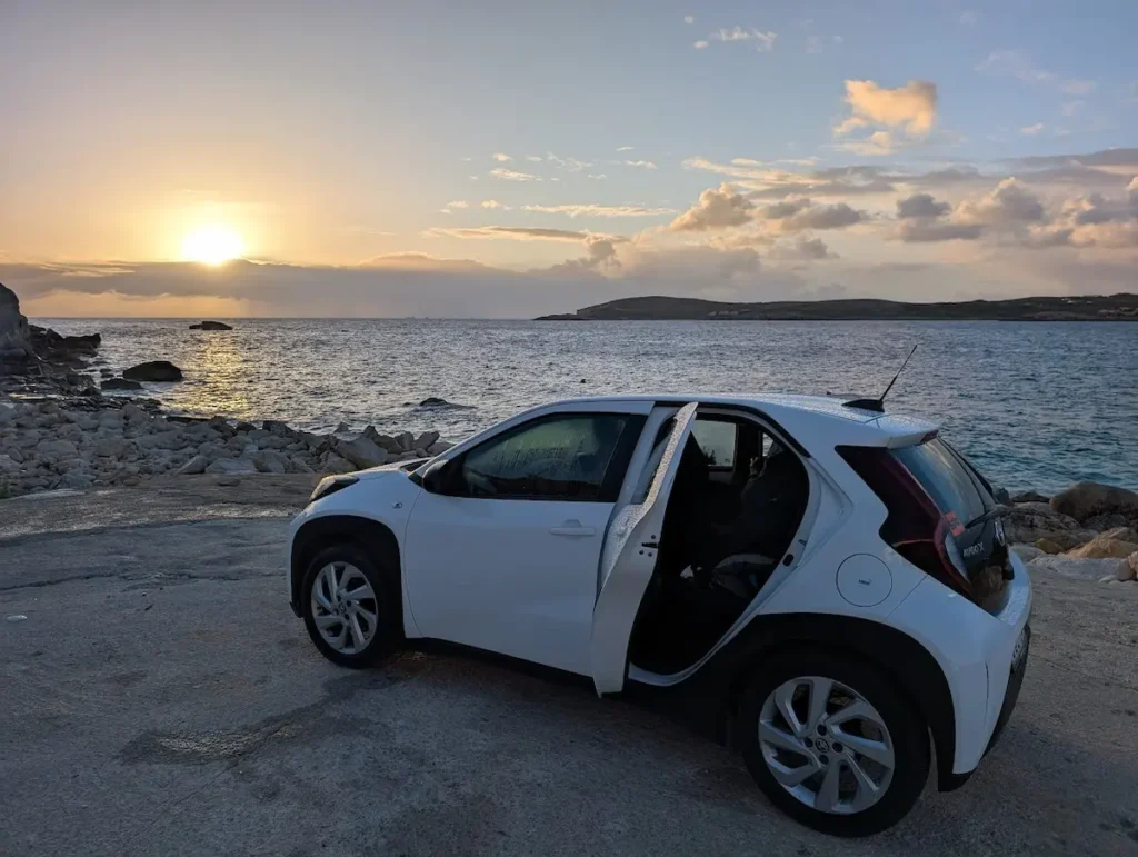 A white Toyota Aygo X rental car parked on a rocky coast in Gozo, Malta, during a vibrant sunset over the Mediterranean Sea.