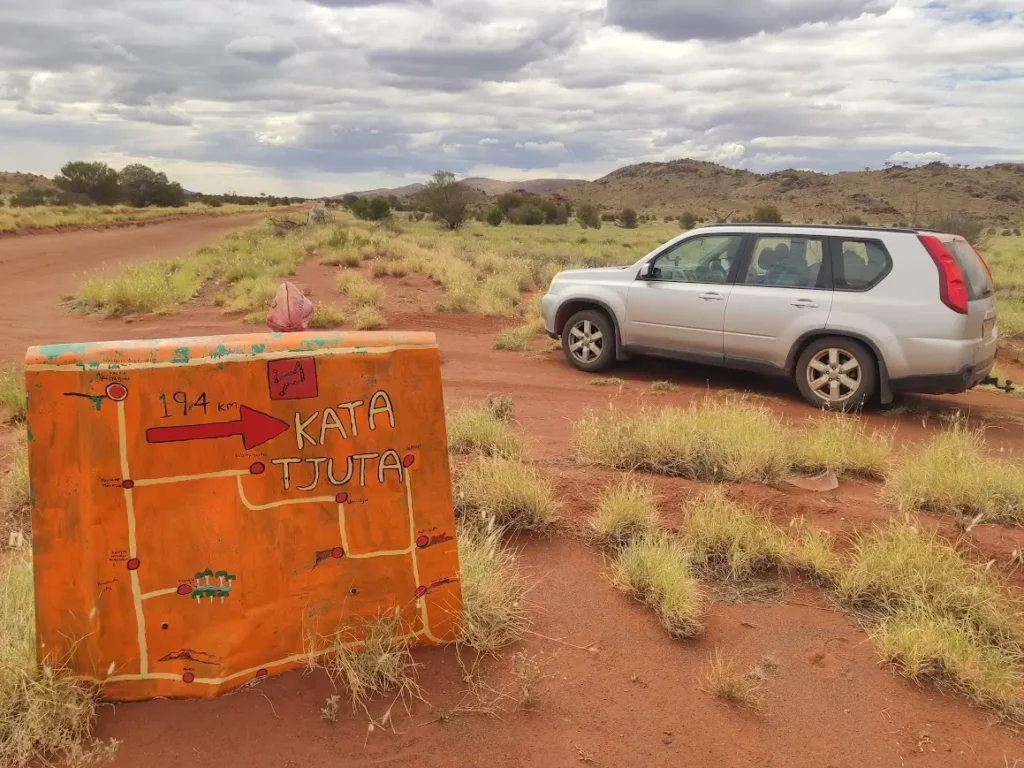 A silver SUV parked on a red dirt road in the Australian Outback next to a hand-painted orange sign pointing to Kata Tjuta.