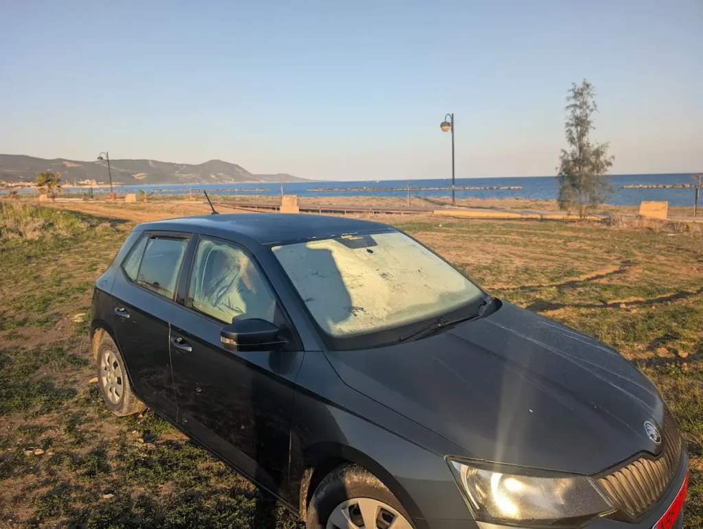 A dark grey Skoda rental car with frost on the windshield parked on a grassy field by the Mediterranean coast in Cyprus during sunrise.