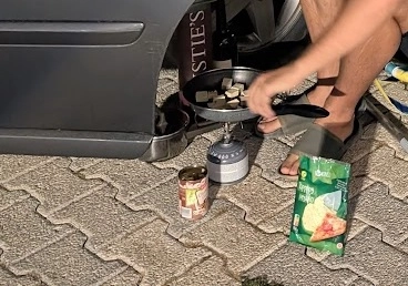 A person cooking tofu in a small frying pan on a SOTO WindMaster camping stove next to a rental car. A bag of chips and a can of food are on the cobblestone ground, illustrating a low-cost mobile kitchen setup.