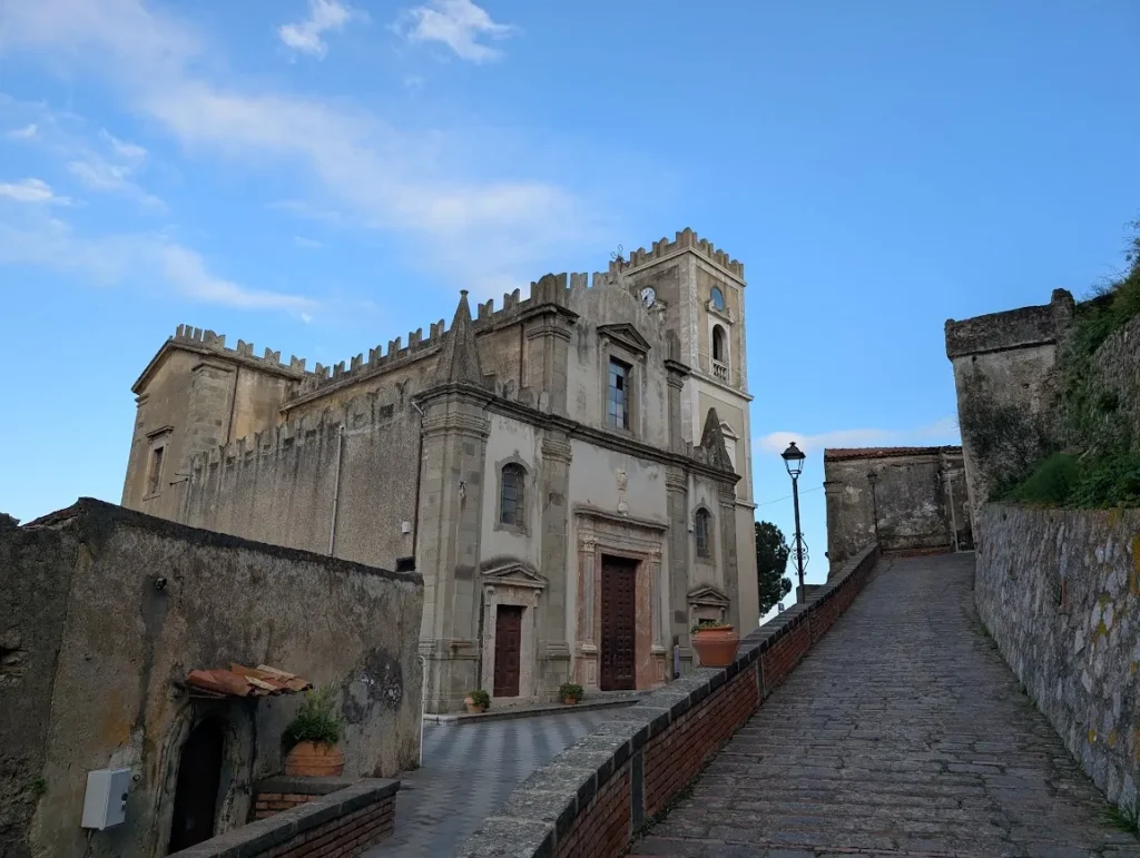 The Chiesa di San Nicolò (aka Santa Lucia) in Savoca, Sicily, a famous Godfather filming location. The stone church has battlement crenellations and a clock tower, positioned next to a steep, cobbled stone ramp.