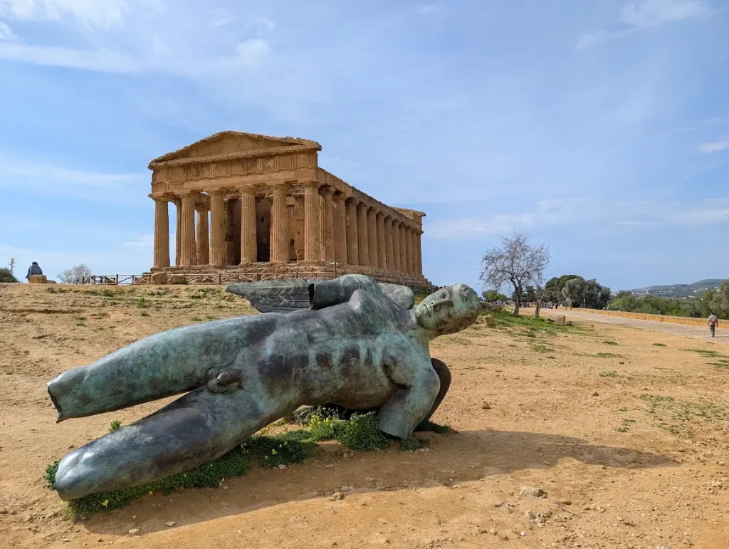 The Temple of Concordia at the Valley of the Temples archeological site in Agrigento, Sicily. A large, green, patinated bronze statue of a fallen Icarus rests on the dusty earth in the foreground. Blue sky.