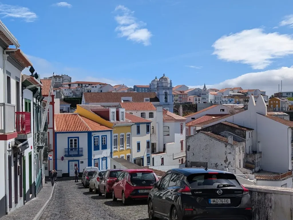 Narrow cobblestone street in Angra do Heroísmo, Terceira, showing traditional colorful architecture and parked cars on a steep incline.