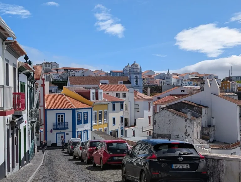 Narrow cobblestone street in Angra do Heroísmo, Terceira, showing traditional colorful architecture and parked cars on a steep incline.