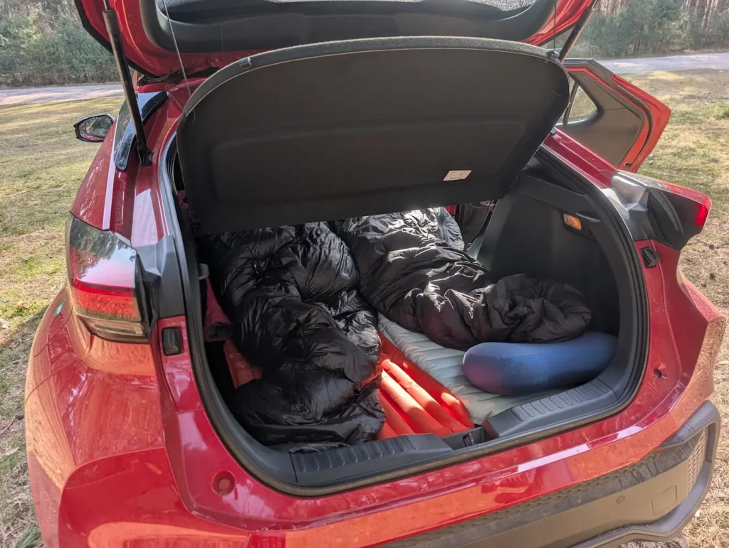 Interior of a red Toyota C-HR parked near the Baltic Sea in Poland, configured as a two-person sleeping area with inflatable pads and sleeping bags.