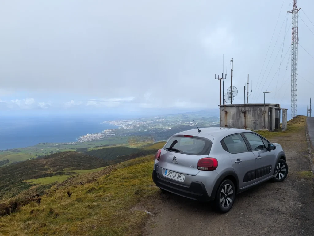 Silver Citroën C3 parked on a hilltop viewpoint in São Miguel, Azores, with coastal town and ocean visible below