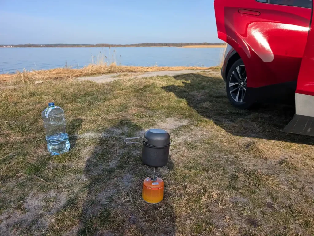 Portable canister stove with cooking pot and a large water bottle on the grass near a lake close to the Baltic Sea in Poland, with the open rear door of a red Toyota C-HR.