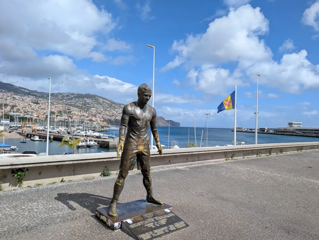 Bronze Cristiano Ronaldo statue on the Funchal waterfront with the marina and Madeira flag behind