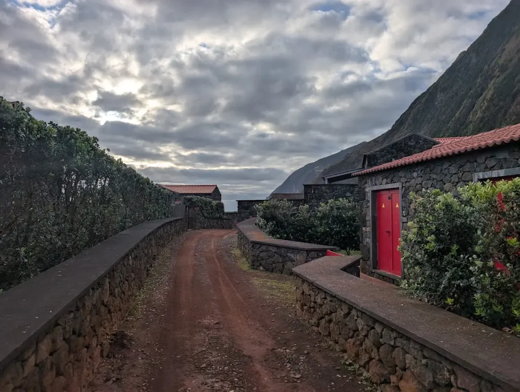 Narrow dirt path between volcanic stone walls and buildings with red doors, cliffs rising behind, Fajã do Belo, São Jorge, Azores