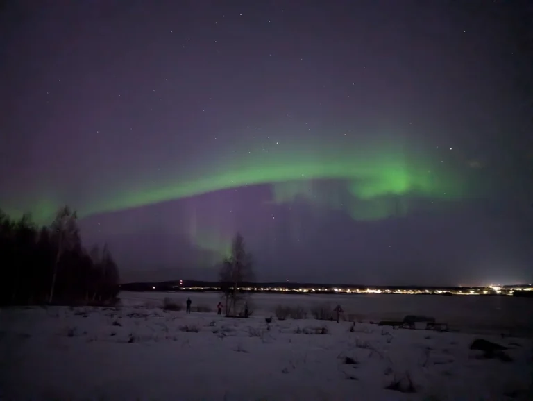 A vibrant display of the Aurora Borealis with green and purple lights arching across the night sky above a snow-covered landscape in Rovaniemi, Finland.