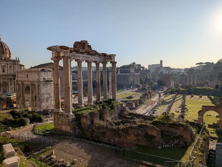 View of the Roman Forum at sunrise with the Temple of Saturn columns in the foreground and the Colosseum visible in the background.
