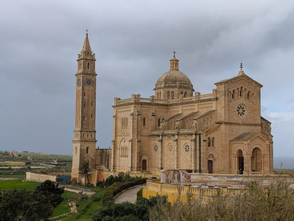 Facade and bell tower of the Basilica of the National Shrine of the Blessed Virgin of Ta' Pinu, Gozo, under a stormy sky.