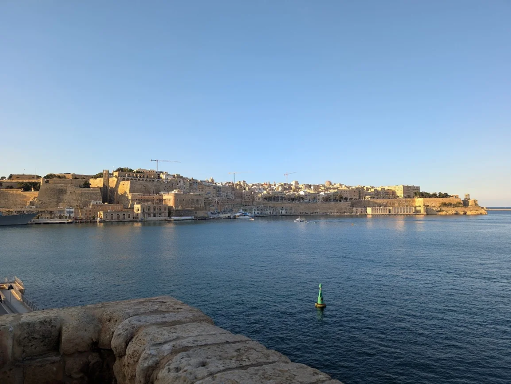 View of Valletta's limestone fortifications and skyline across the Grand Harbour, seen from Senglea at golden hour.