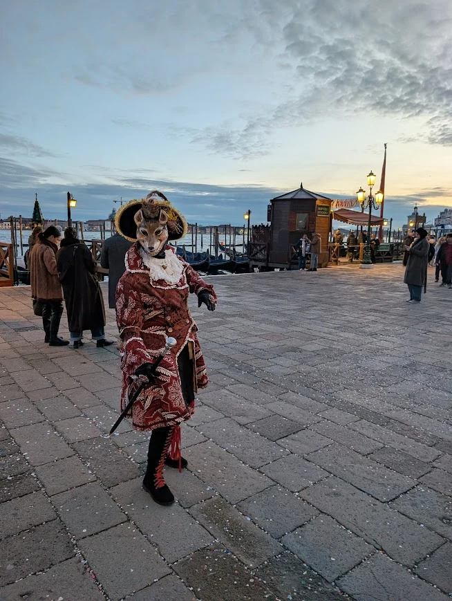 Person in elaborate Venice Carnival costume and fox mask on the waterfront at dusk