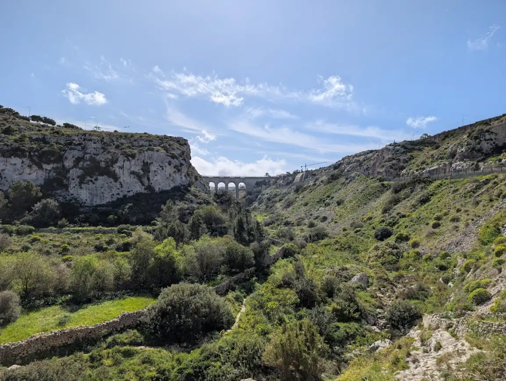 Green valley with dry-stone walls and dense scrub vegetation in central Malta, with a Roman-style aqueduct bridge visible in the background.