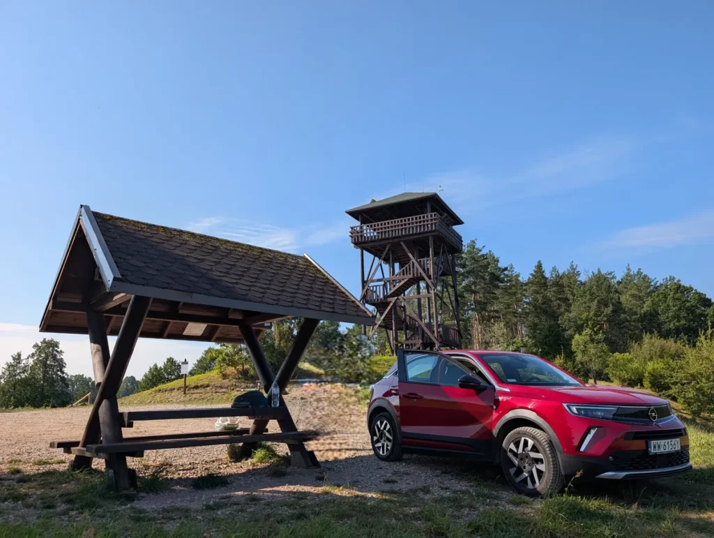 Red Opel Mokka parked at a free overnight spot in Poland with a wooden watchtower in the background