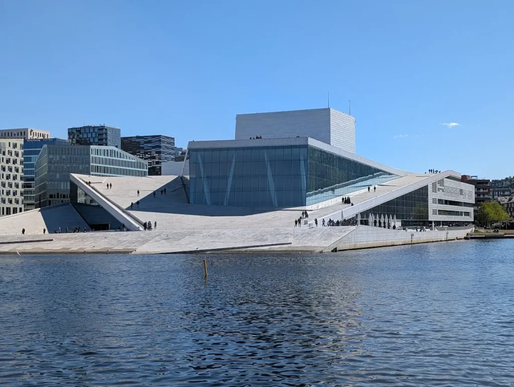 Oslo Opera House seen from the waterfront, with people walking on its sloped marble roof