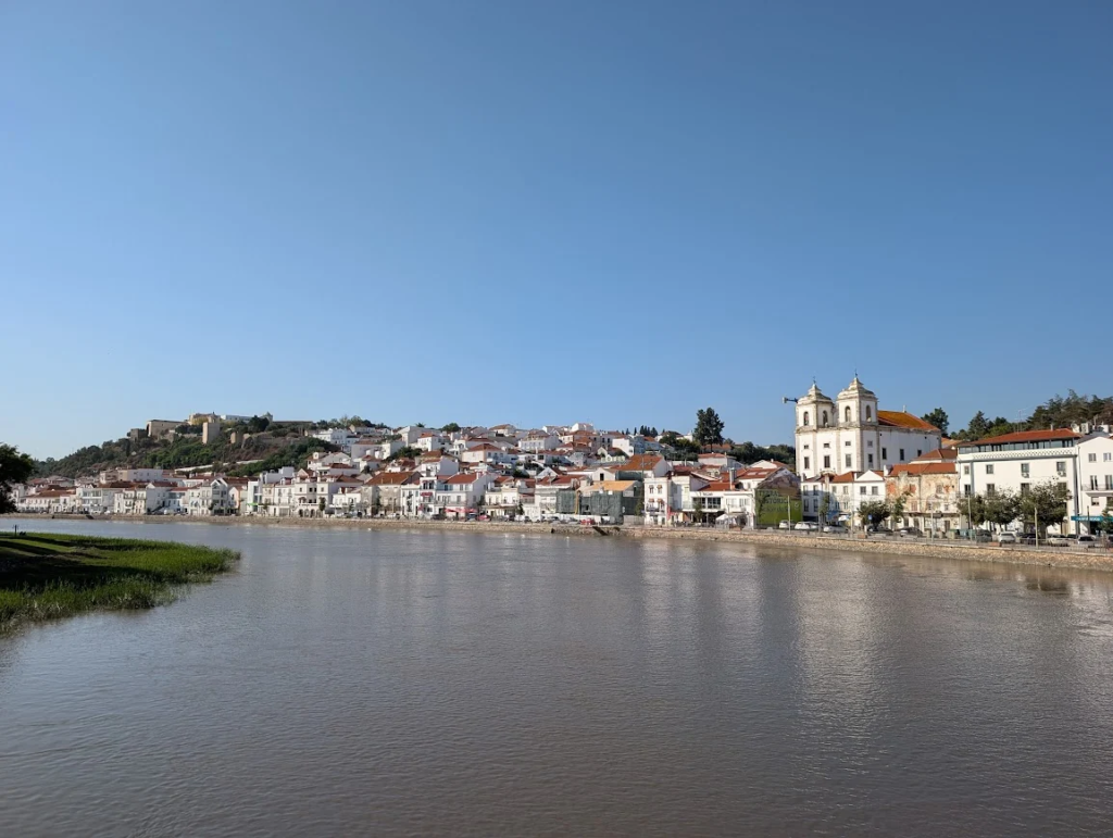 Alcácer do Sal waterfront viewed from across the Sado river, with the castle on the hill and whitewashed buildings along the bank