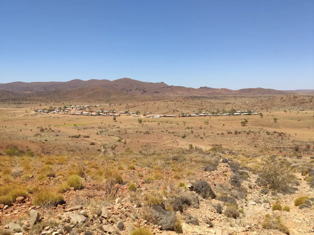 View of Pipalyatjara community from a nearby hill, with houses scattered across the desert landscape and mountains in the background.