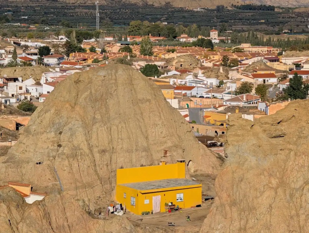 Yellow house built into badlands rock formations with the town of Guadix visible behind, Spain