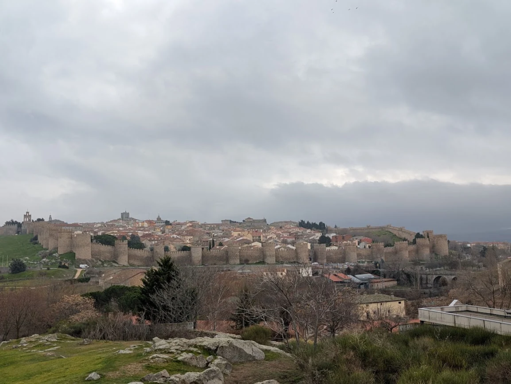 Medieval city walls of Ávila, Spain, seen from a hillside lookout under heavy grey clouds on a rainy December day