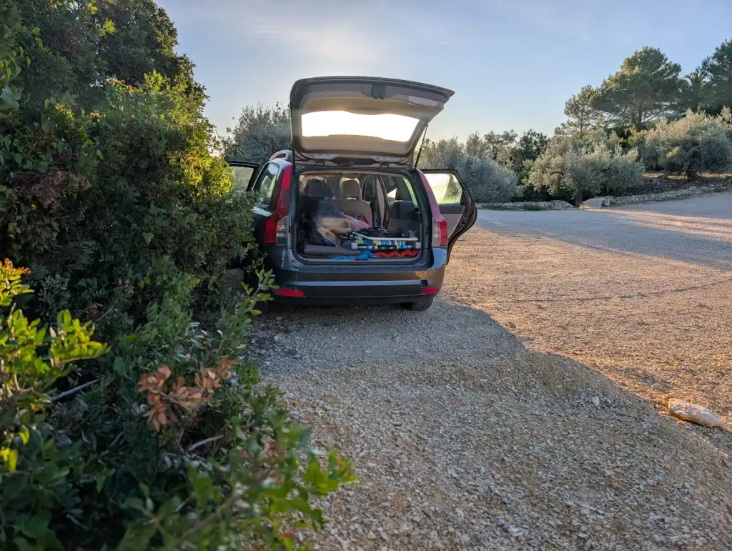 Car with rear hatch open at a coastal camper parking spot in Peñíscola at sunrise, sleeping setup visible in the trunk