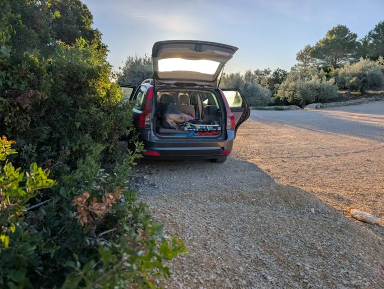 Car with rear hatch open at a coastal camper parking spot in Peñíscola at sunrise, sleeping setup visible in the trunk