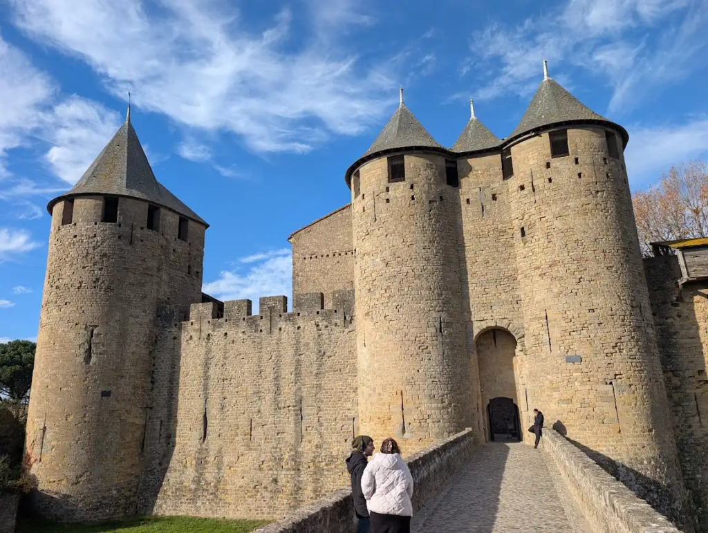 Entrance gate of the Château Comtal in Carcassonne with round stone towers and conical roofs, and visitors crossing the stone bridge