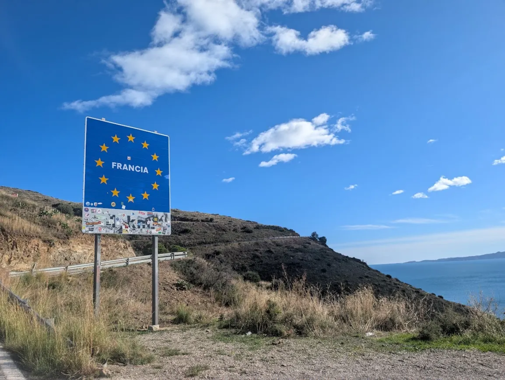 Blue EU Francia border sign covered in stickers on the coastal road between Catalonia and southern France, with the Mediterranean behind it