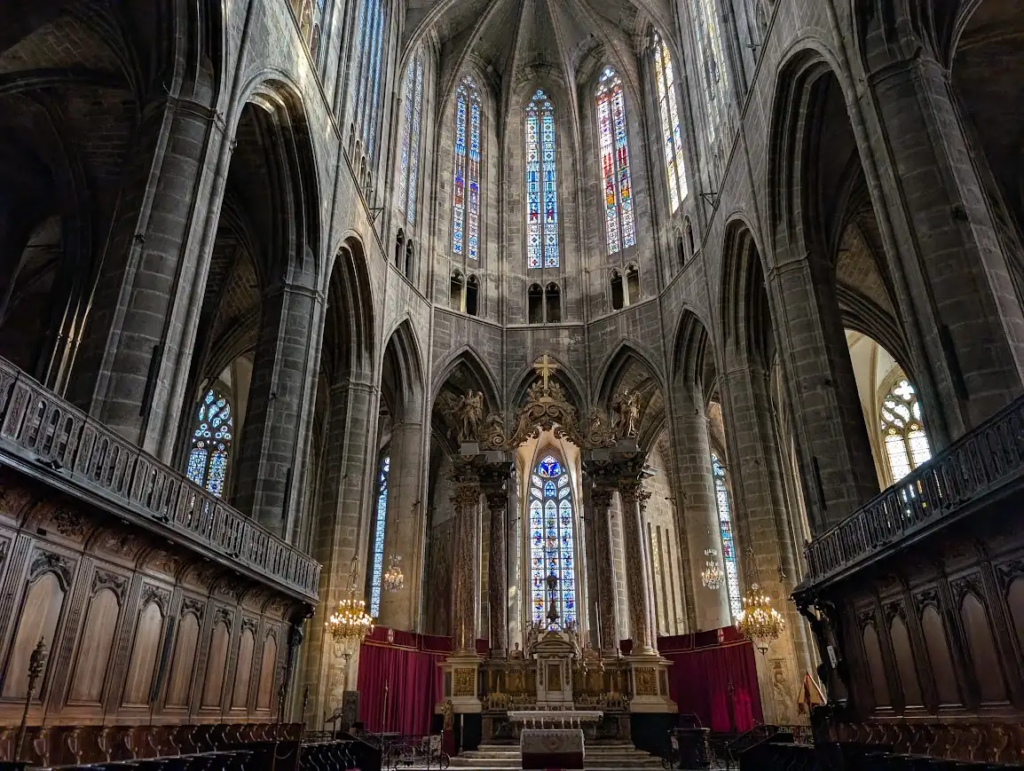 Interior of Narbonne cathedral in High Gothic style, with tall stained glass windows above the altar and carved wooden choir stalls