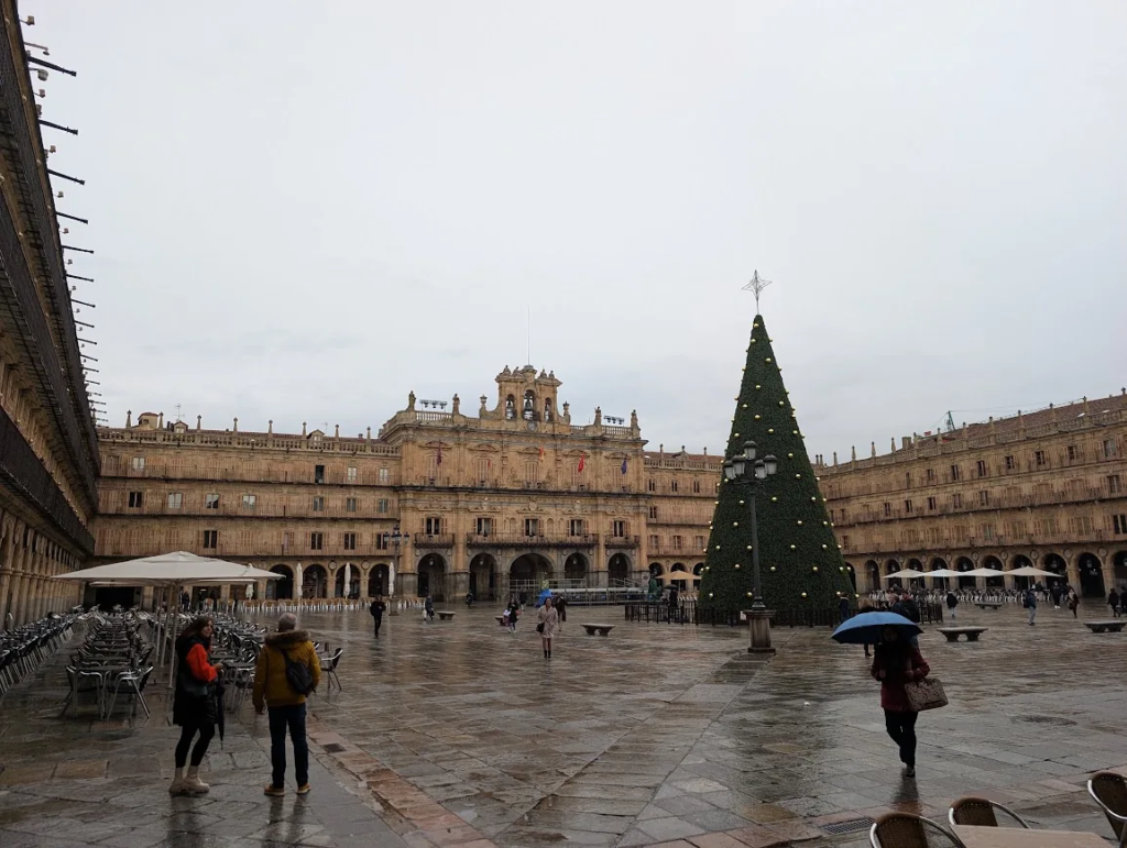 Plaza Mayor in Salamanca, Spain, with wet cobblestones, a large Christmas tree and empty café chairs on a rainy December day