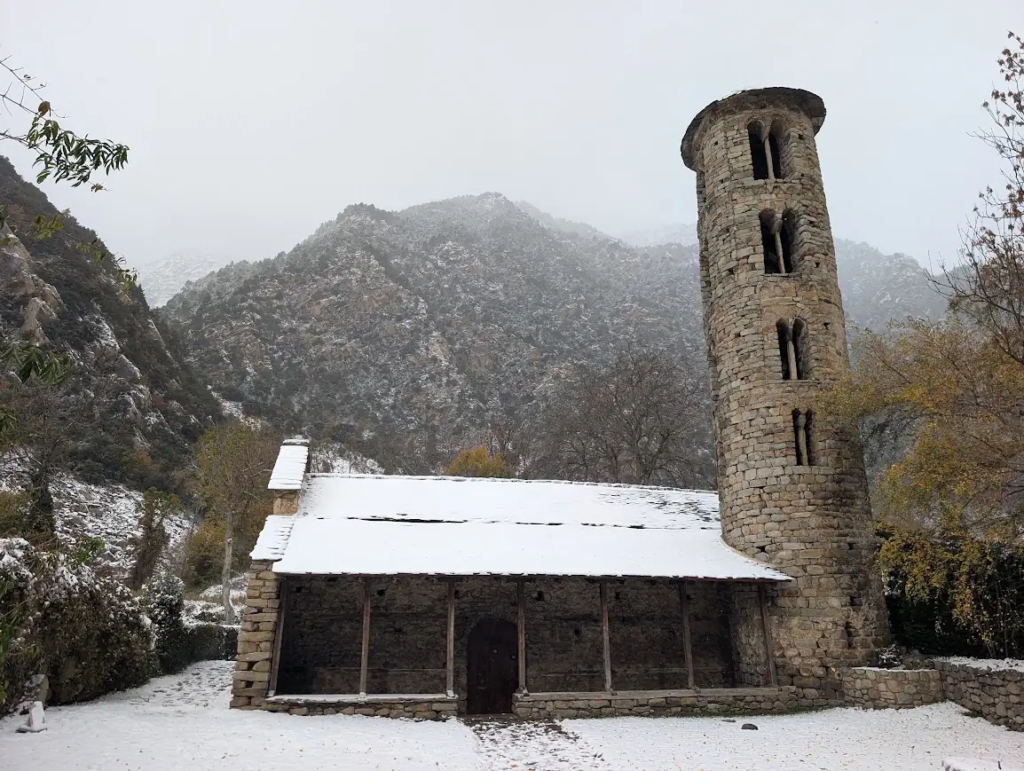 Romanesque church of Santa Coloma in Andorra with a round stone bell tower and snow-covered roof, in front of snowy mountains