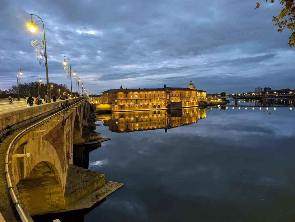 Garonne river in Toulouse at dusk with an illuminated brick building reflected in the water and a stone bridge lined with decorative streetlights