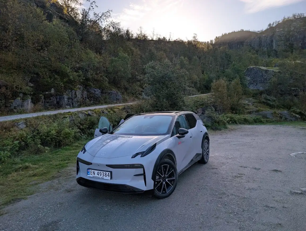 White Zeekr X electric SUV with Norwegian plates parked on a gravel area below a rocky, tree-covered hillside, driver door open, morning sun behind the ridge.