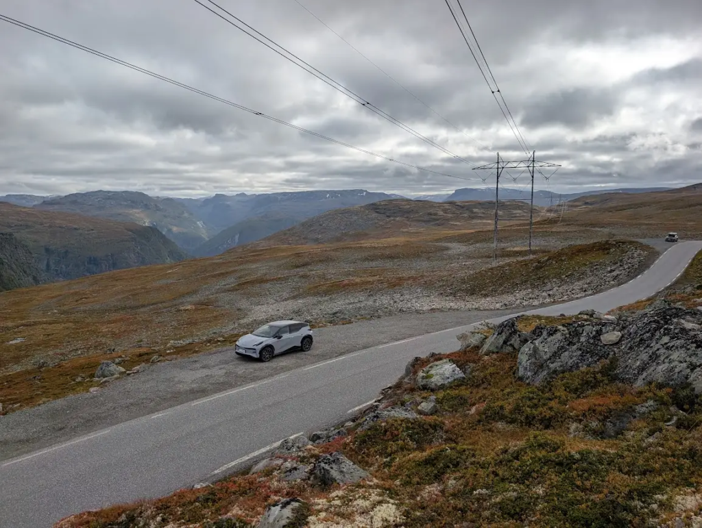 White electric rental car parked at a pull-off on the Aurlandsfjellet mountain road, with power lines and a bare plateau under heavy cloud
