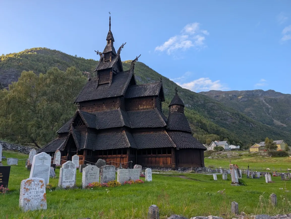 Borgund Stave Church with its tiered dark wooden roofs and dragon heads, surrounded by a churchyard and green mountains under a blue sky