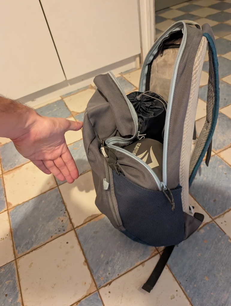 Open gray Quechua Arpenaz daypack loaded with sleeping gear and kit, with a hand extended next to it for size scale.