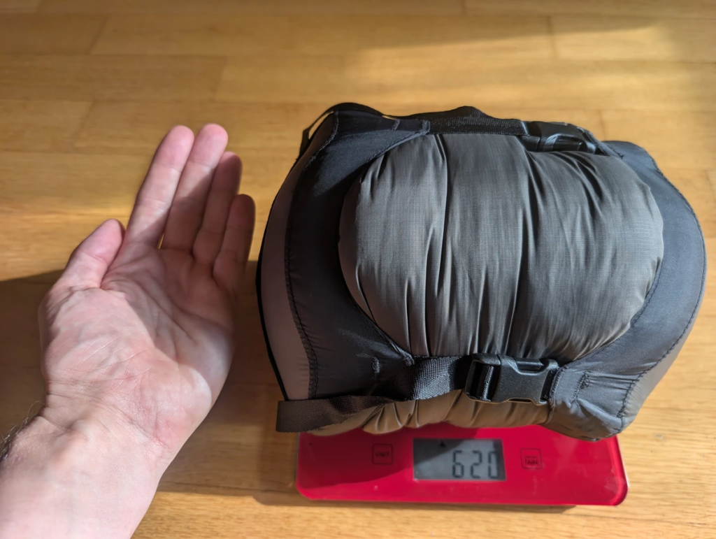 Sea to Summit Spark -1C sleeping bag compressed in a gray stuff sack sitting on a red digital scale reading 620 grams, with a hand alongside it for size comparison.