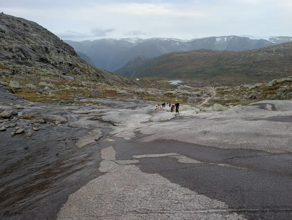 Hikers on wet bare granite slabs on the Trolltunga trail, with snow-dusted mountains in the distance