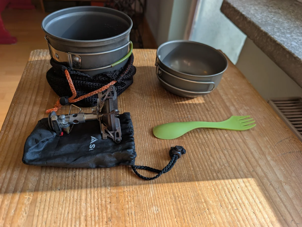 Soto WindMaster stove with black pouch, green spork, and a 1,000ml nesting aluminum pot set arranged on a wooden table.