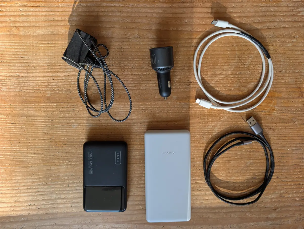 Close-up layout of ultralight tech accessories including INIU fast charge power bank, gray Xiaomi power bank, JOYROOM 120W car charger, Nitecore headlamp, and USB cables on a wood surface.