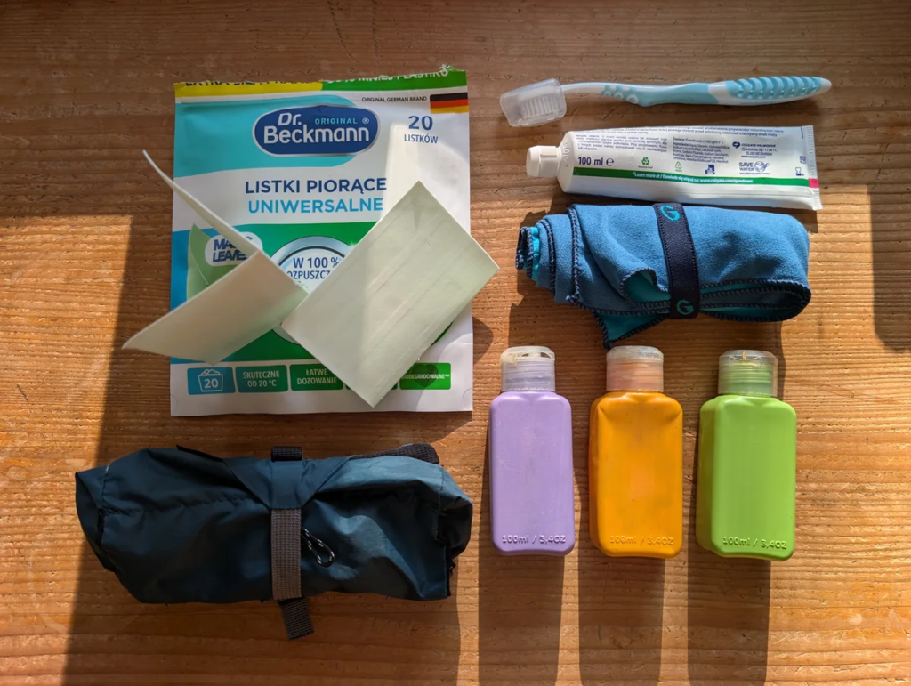 Minimalist hygiene layout featuring Dr. Beckmann universal laundry sheets, a couple of 100ml travel bottles, toothbrush, toothpaste, rolled microfiber towel, and zippered pouch on a wood surface.